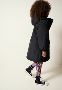 Sort hættefrakke med elastiske manchetter, parret med farverigt splattemønster leggings og sorte højtop sneakers.