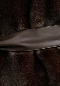 Brązowe futro o miękkiej fakturze, połączone z gładką brązową skórzaną opaską, tworzącą warstwowy wzór. Skórzany akcent ma prostokątny i płaski kształt.