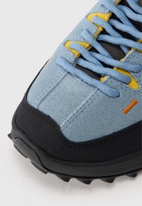 Ανοιχτό μπλε σουέντ αθλητικό παπούτσι με μαύρη ελαστική σόλα, μπλε κορδόνια και κίτρινες και πορτοκαλί λεπτομέρειες. Υφή επιφάνειας με αντιθετικές ραφές.