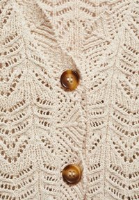 Pletený cardigan ve světle béžové barvě s motivem listů, texturovanou tkaninou a velkým hnědým knoflíkem na zapínání vpředu.