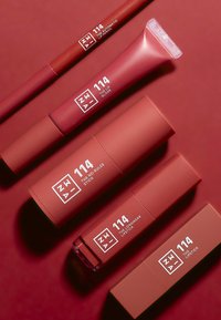 Five red lip products arranged on a matching red background: lip pencil, gloss, longwear stick, and two lipsticks, all with white labels.