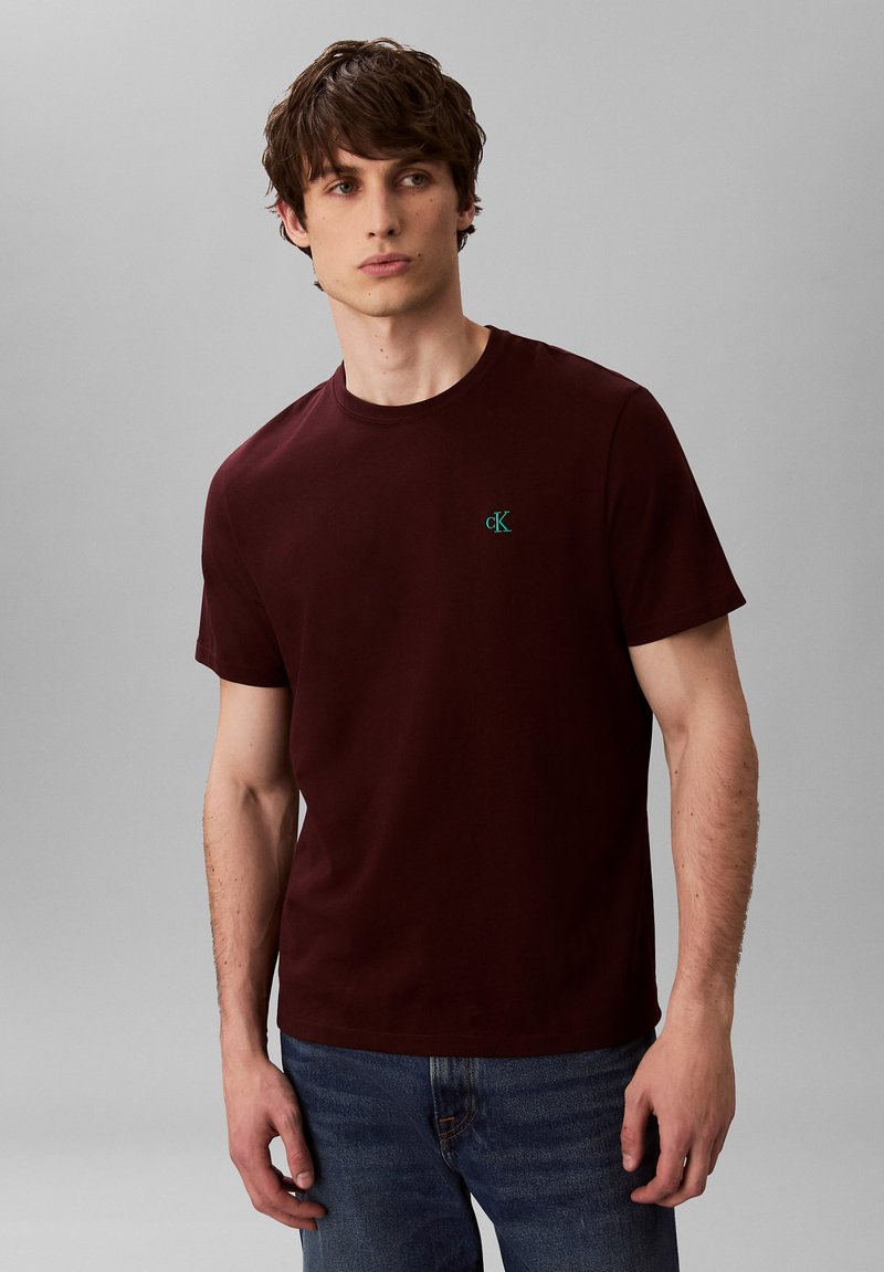 Ung mand iført en mørkerød kortærmet t-shirt med lille grønblåt "cK"-logo på brystet, kombineret med blå jeans, stående foran en ensfarvet grå baggrund.