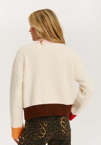 Sweter dzianinowy w kolorze kremowym z pomarańczowymi, brązowymi i czerwonymi akcentami. Posiada teksturowany wzór w zygzak na jednym rękawie oraz krótki krój.