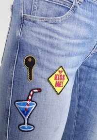 Svetle modré džíny s třemi barevnými vyšitými nášivkami: klíčem, žlutým označením s nápisem "KISS ME!" a koktejlovou sklenicí se pruhovanou brčkem.