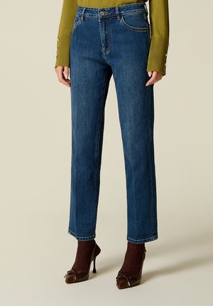 Person trägt blaue Straight-Leg-Jeans, olivgrünen Pullover mit Knöpfen und langen Ärmeln, braune High Heels und braune Socken.