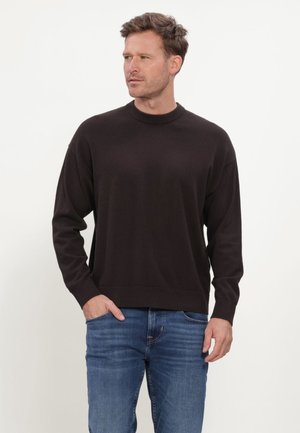 Uomo con un maglione nero semplice a maniche lunghe e jeans blu, in piedi con una mano in tasca, che guarda a sinistra.
