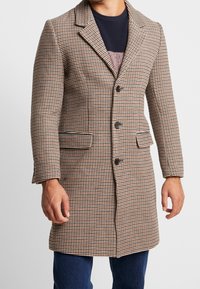 Hombre vistiendo un abrigo de cuadros marrón, negro y blanco con botones, bolsillos de solapa, suéter de punto oscuro y jeans azules contra un fondo simple.