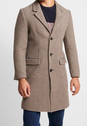 Homme portant un manteau à carreaux marron, noir et blanc avec des boutons, des poches à rabat, un pull en tricot foncé et un jean bleu devant un fond uni.