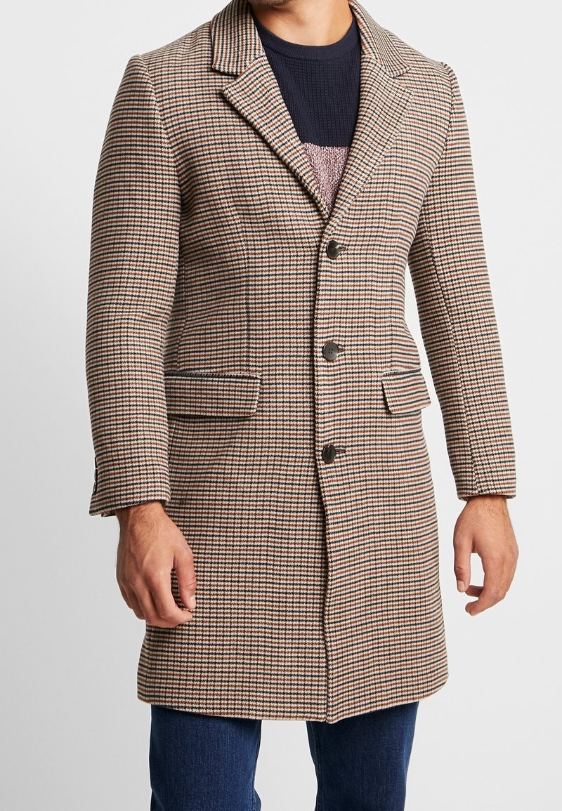 Hombre vistiendo un abrigo de cuadros marrón, negro y blanco con botones, bolsillos de solapa, suéter de punto oscuro y jeans azules contra un fondo simple.