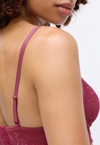 Primo piano di una spalla e parte alta della schiena di una donna che indossa un reggiseno di pizzo bordeaux con spalline regolabili, su uno sfondo bianco.