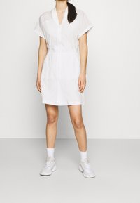 Kobieta ubrana w białą sukienkę do kolan z krótkimi rękawami oraz białe buty sportowe z białymi skarpetkami na jednolitym tle.