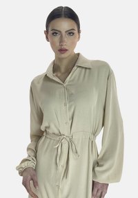 Robe chemise beige à manches longues avec un devant boutonné, une taille ajustable à nouer et un tissu doux et texturé. Elle présente un col classique et des poignets froncés.