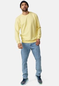 Gul sweatshirt med rund hals og ribbede manchetter, parret med lyseblå jeans og grå træningssko med blå og grønne accenter.