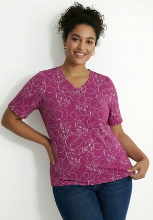Jeune femme souriante, portant un t-shirt à col en V magenta avec des dessins abstraits blancs en lignes, et un jean bleu, debout contre un mur uni.