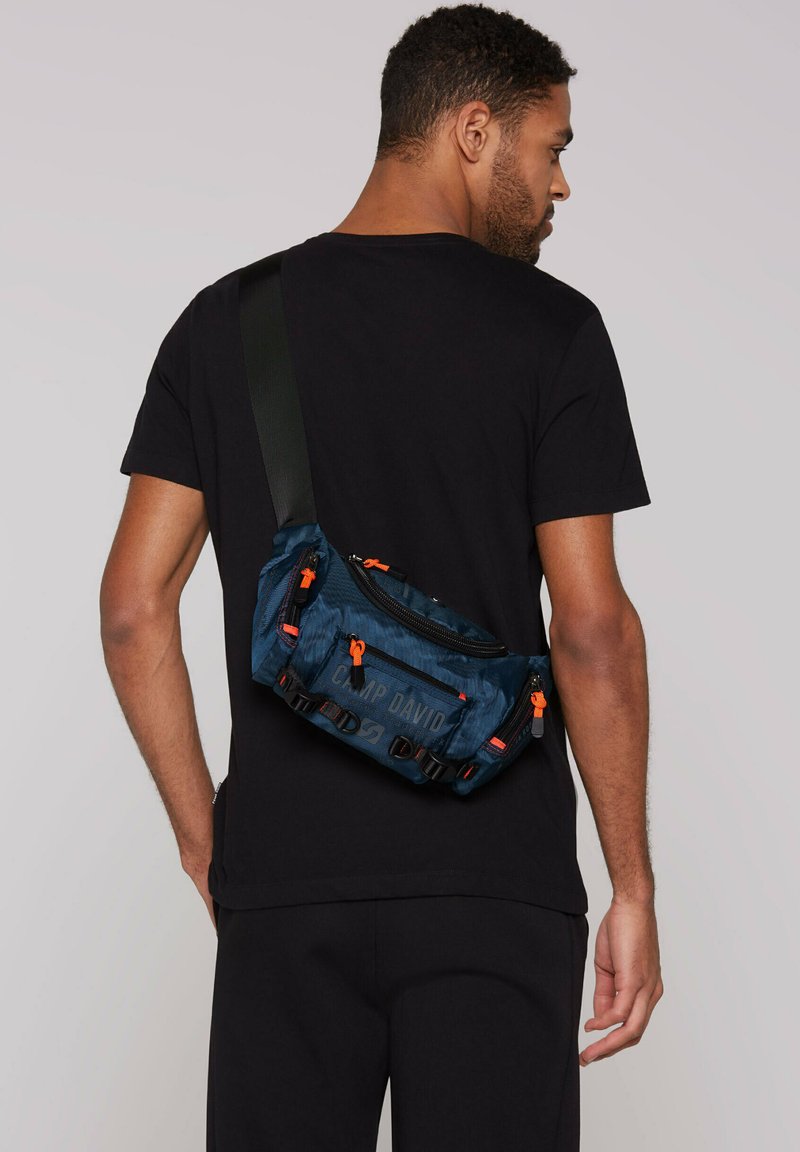 Blaues Nylon-Slingbag mit schwarzen Akzenten, orangen Reißverschluss-Zippern und einem Logo. Verfügt über mehrere Fächer und einen verstellbaren Schultergurt.