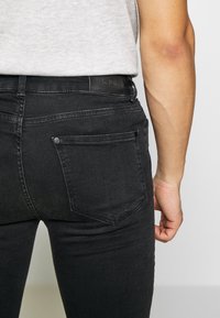 Vaqueros de mezclilla negra con corte ajustado, costuras en contraste, cinco bolsillos y un parche de cuero en la cintura. Textura suave, diseño minimalista.