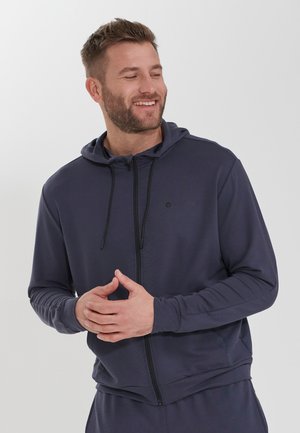 Sweat-shirt zippé navy en tissu lisse, avec une capuche à cordon, des poches et un logo subtil sur la poitrine, offrant une coupe décontractée.
