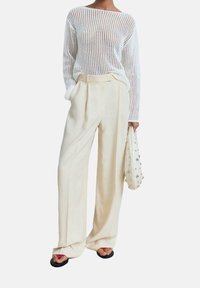 Pull en tricot blanc avec un col large, associé à un pantalon large plissé de couleur crème. Le mannequin tient un sac beige avec des accents métalliques.