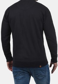 Musta svetari, jossa pyöreä pääntie, resorit hihansuissa ja helmassa. Selässä pieni logotarra vyötärön yläpuolella.