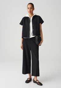 Black cropped jacket with short sleeves and front buttons, worn over a white hoodie, paired with black wide-leg trousers and woven shoes.
