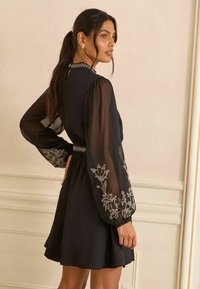 Fekete ruha átlátszó hosszú ujjakkal, díszített virágos hímzéssel, karcsúsított derékkal és bővülő szoknyával. Magas nyakkal és hátul kivágással rendelkezik.