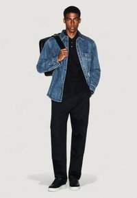 Giacca di jeans sopra una polo nera, pantaloni cargo neri e sneakers nere. Zaino indossato su una spalla; la giacca presenta tasche frontali.