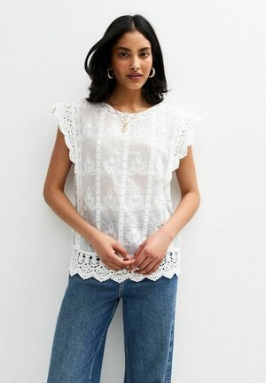 Haut en dentelle blanche avec des manches festonnées, une broderie complexe et un ourlet en crochet. Associé à un pantalon en denim bleu.