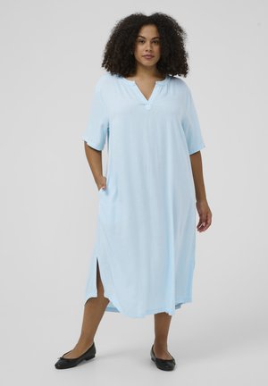 Kvinde med krøllet hår iført en lyseblå midi-kjole med sideslidser og sorte flade sko, stående mod en ensfarvet baggrund.