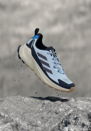 Světle modrá běžecká obuv Adidas Terrex s černými pruhy a hnědou podrážkou zavěšená nad skalnatým terénem s částicemi hlíny.