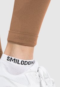 Bruine legging van rekbare stof met een omgeslagen onderkant. Gecombineerd met witte sneakers met een zwart logo op de sok.