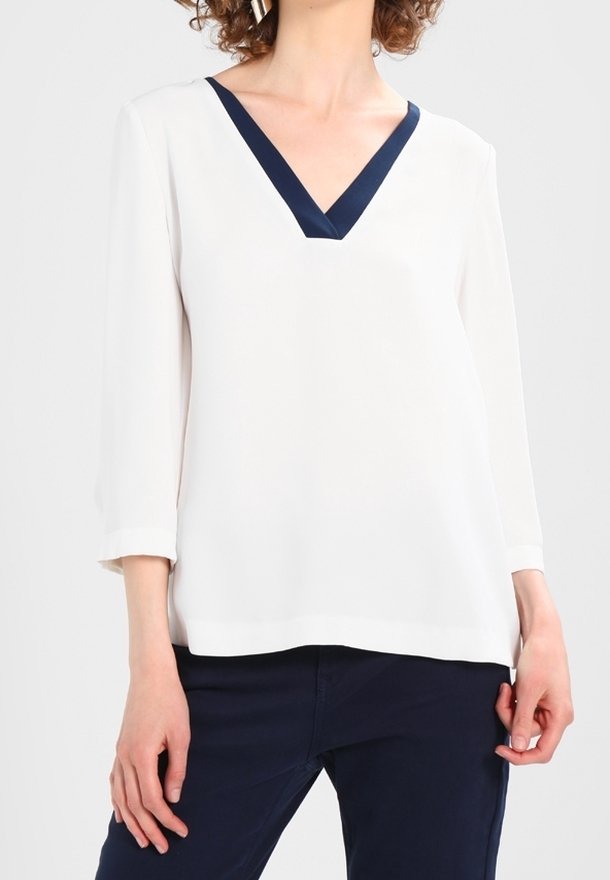 Femme portant un blouse blanche à col en V avec des finitions marine et des manches trois-quarts, associée à un pantalon marine, se tenant devant un fond uni.