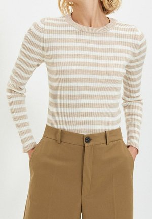 Femme portant un haut côtelé beige et blanc à rayures horizontales, manches longues, rentré dans un pantalon camel marron taille haute, les mains dans les poches.