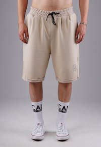 Beige cotton shorts with an elastic waistband and drawstring, featuring side pockets and a logo detail on the left leg. Worn with white socks.