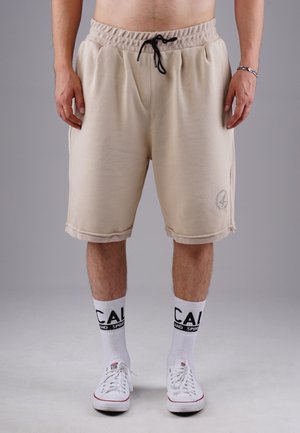 Beige cotton shorts with an elastic waistband and drawstring, featuring side pockets and a logo detail on the left leg. Worn with white socks.