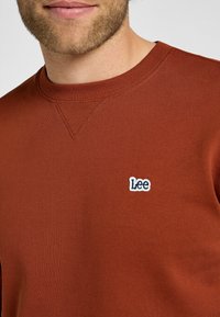 Felpa color ruggine con collo a giro e texture a coste. Presenta un piccolo logo "Lee" ricamato in bianco sul petto.