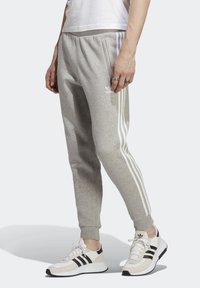 Pantalones de chándal grises hechos de una tela suave, con tres rayas blancas a lo largo de los lados y un pequeño logo blanco en el muslo izquierdo.