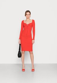 Vestido vermelho canelado com decote quadrado, mangas longas e detalhe de cordão na frente. Combinado com sandálias vermelhas de salto aberto e uma bolsa preta.