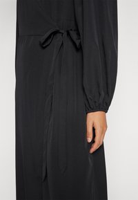 Svart klänning med långa ärmar och knytning i midjan. Mjukt tyg med en slät yta och något rynkade ärmslut. Sidofickor synliga.