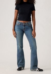 Femme portant un haut court noir à manches courtes, jean évasé bleu, baskets noires et blanches, debout devant un fond clair uni.