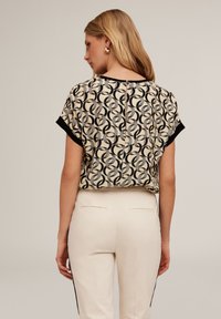 Kortärmad blus i krämfärgad med ett cirkulärt svart och grått mönster, med knäppning bak och svarta dekorationer, bärs med ljusa byxor.