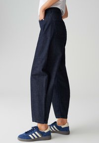 Pantalon en jean foncé avec une coupe droite et ample, taille haute, poches latérales et arrière, associé à des baskets bleues et blanches.