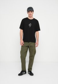 Camiseta negra de algodón con un diseño floral blanco, combinada con pantalones cargo verde oliva con múltiples bolsillos y zapatillas negras.