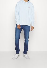 Sudadera con capucha azul claro con bolsillo delantero, capucha con cordón y pequeño logo en el pecho, combinada con pantalones vaqueros azules y zapatillas blancas.