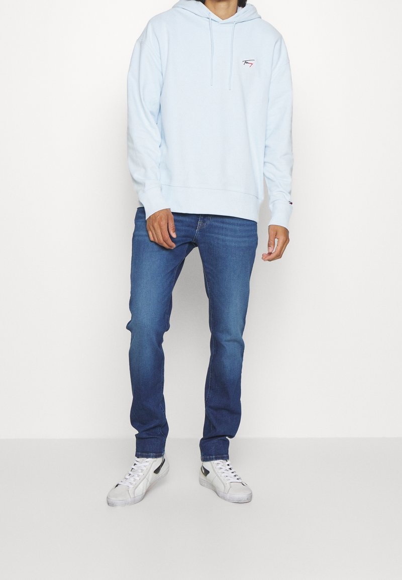 Sudadera con capucha azul claro con bolsillo delantero, capucha con cordón y pequeño logo en el pecho, combinada con pantalones vaqueros azules y zapatillas blancas.