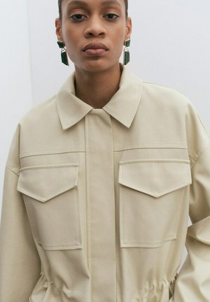 Femme portant une veste beige à col avec de grandes poches avant et des boucles d'oreilles pendantes géométriques vertes, debout devant un fond clair uni.