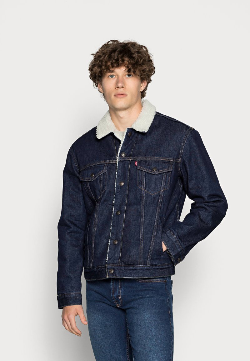 Mörk denimjacka med en vit foderkrage, framknappar och två bröstfickor. Designen inkluderar synliga sömmar och en figurnära silhuett.