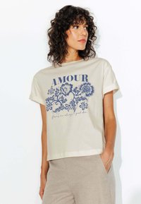 T-shirt en coton crème avec des manches roulées, ornée d'un motif floral bleu et du texte "AMOUR" au-dessus de la phrase "les fleurs sont toujours une bonne idée."