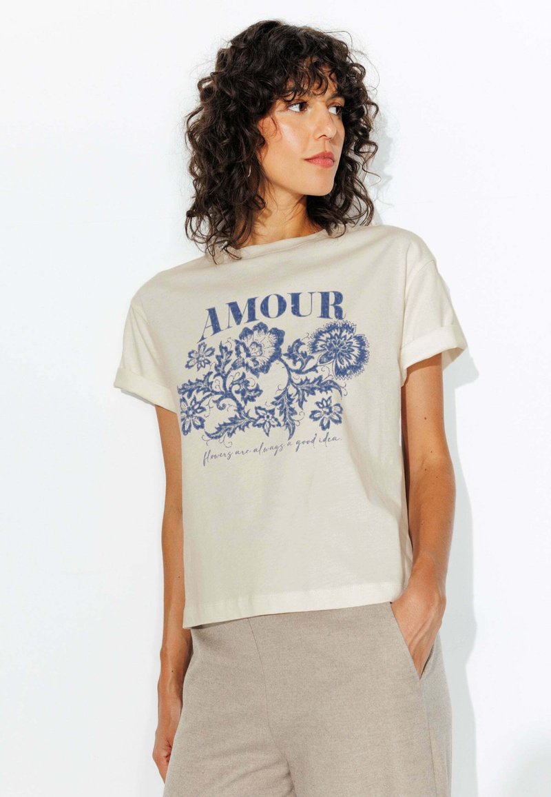 T-shirt en coton crème avec des manches roulées, ornée d'un motif floral bleu et du texte "AMOUR" au-dessus de la phrase "les fleurs sont toujours une bonne idée."