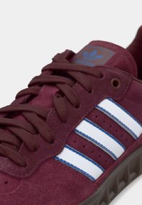 Burgundyröda mockasneakers med vita och blå ränder, med platta snören och en blå Adidas-logotyp på tungan.
