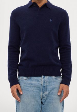 Homme portant un pull polo bleu marine à manches longues et un jean bleu clair, les mains détendues sur les côtés.